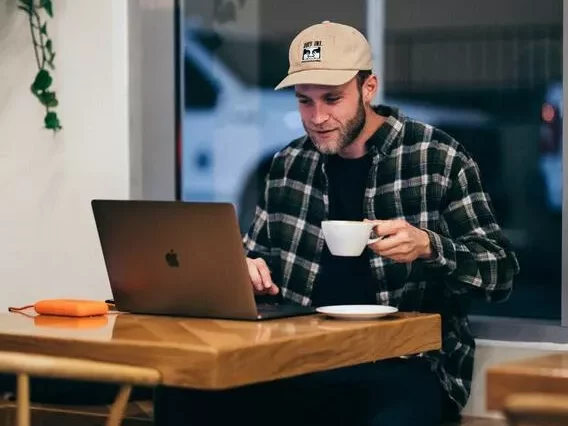 Ein Mann sitzt mit einem Laptop und einer Tasse Kaffee an einem Tisch.