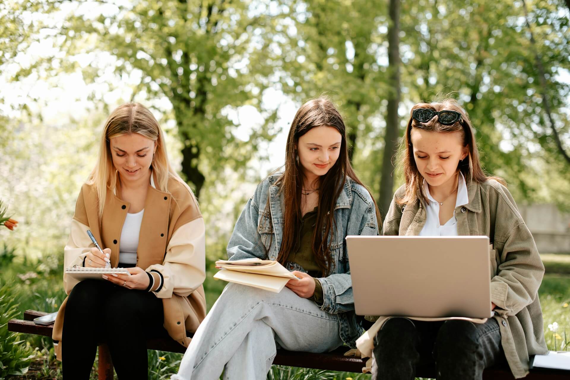 3 Schülerinnen sitzen zusammen auf einer Parkbank und lernen am Laptop