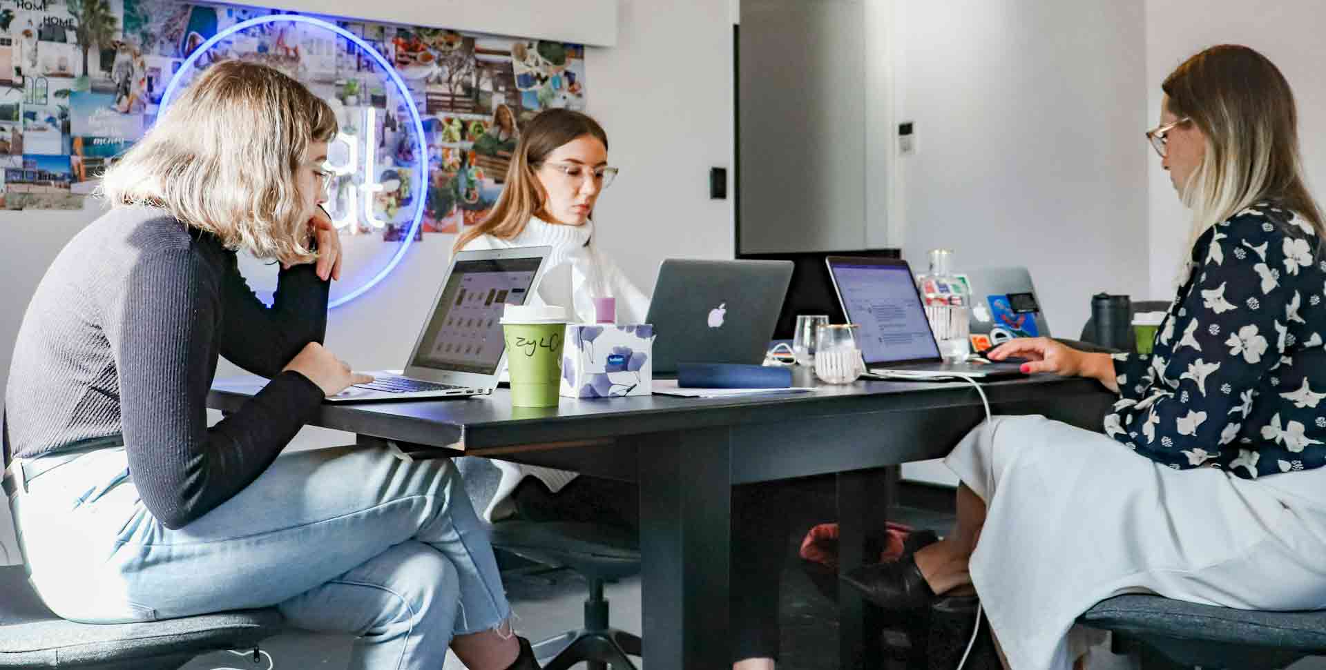 Drei junge Frauen arbeiten in modernem Startup-Büro an Laptops mit Neon-Schild und Mood-Board.