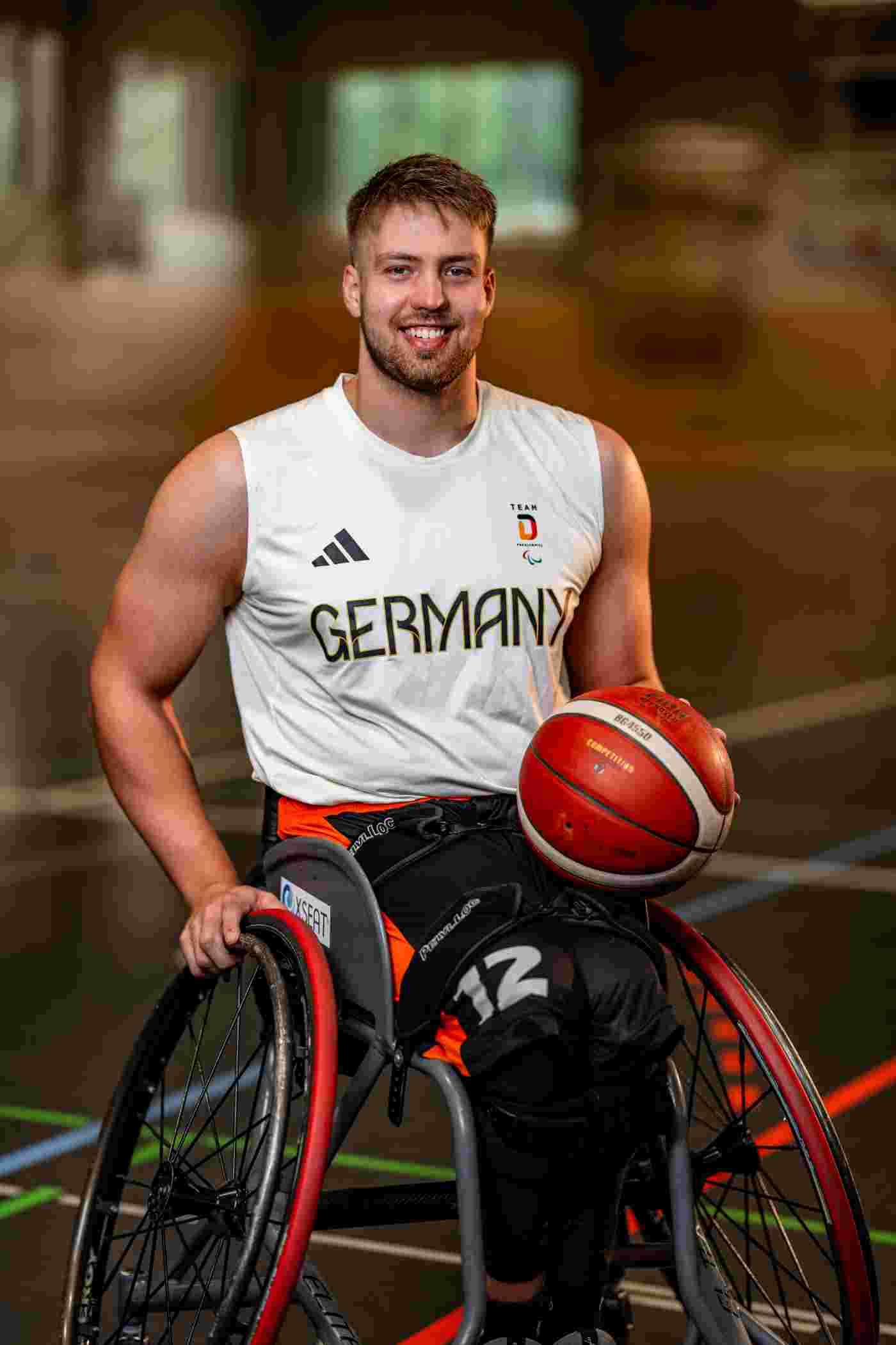 Alexander Budde in weißem Team D Adidas-Trikot hält roten Basketball auf seinem Sportrollstuhl
