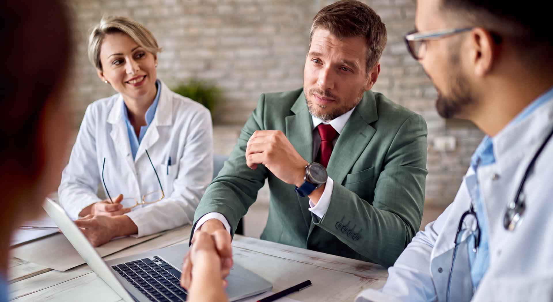 Geschäftstreffen im Gesundheitswesen: Ein Manager in grünem Anzug diskutiert mit medizinischem Fachpersonal an einem Konferenztisch mit Laptop