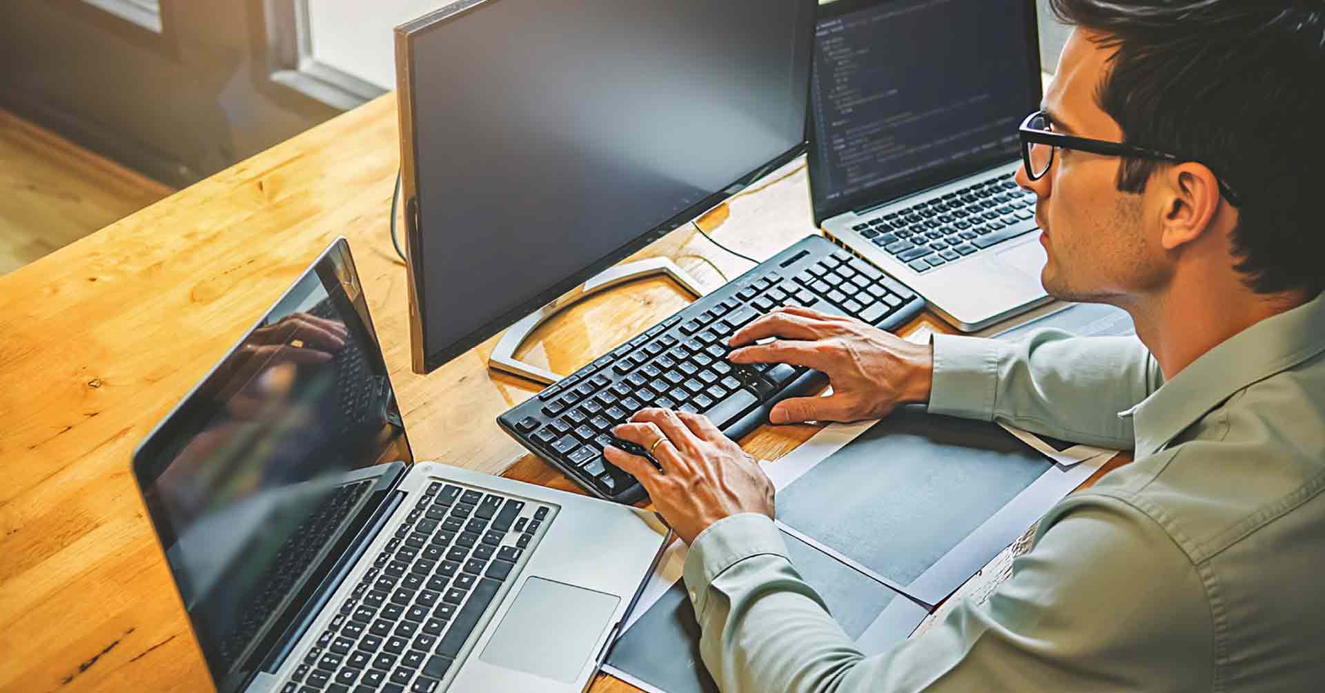 Softwareentwickler mit Brille arbeitet an mehreren Bildschirmen und Laptops an einem Holzschreibtisch.