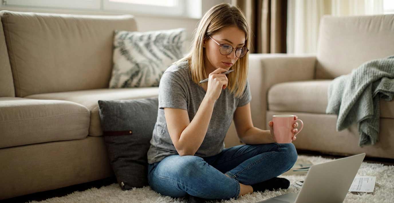 Frau mit Brille sitzt auf dem Boden vor einem Laptop, hält einen rosa Becher und arbeitet konzentriert an ihrem Computer.