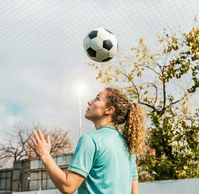 Junge Fußballspielerin in türkisem Trikot macht einen Kopfball auf einem Außenplatz mit Netz und Bäumen im Hintergrund